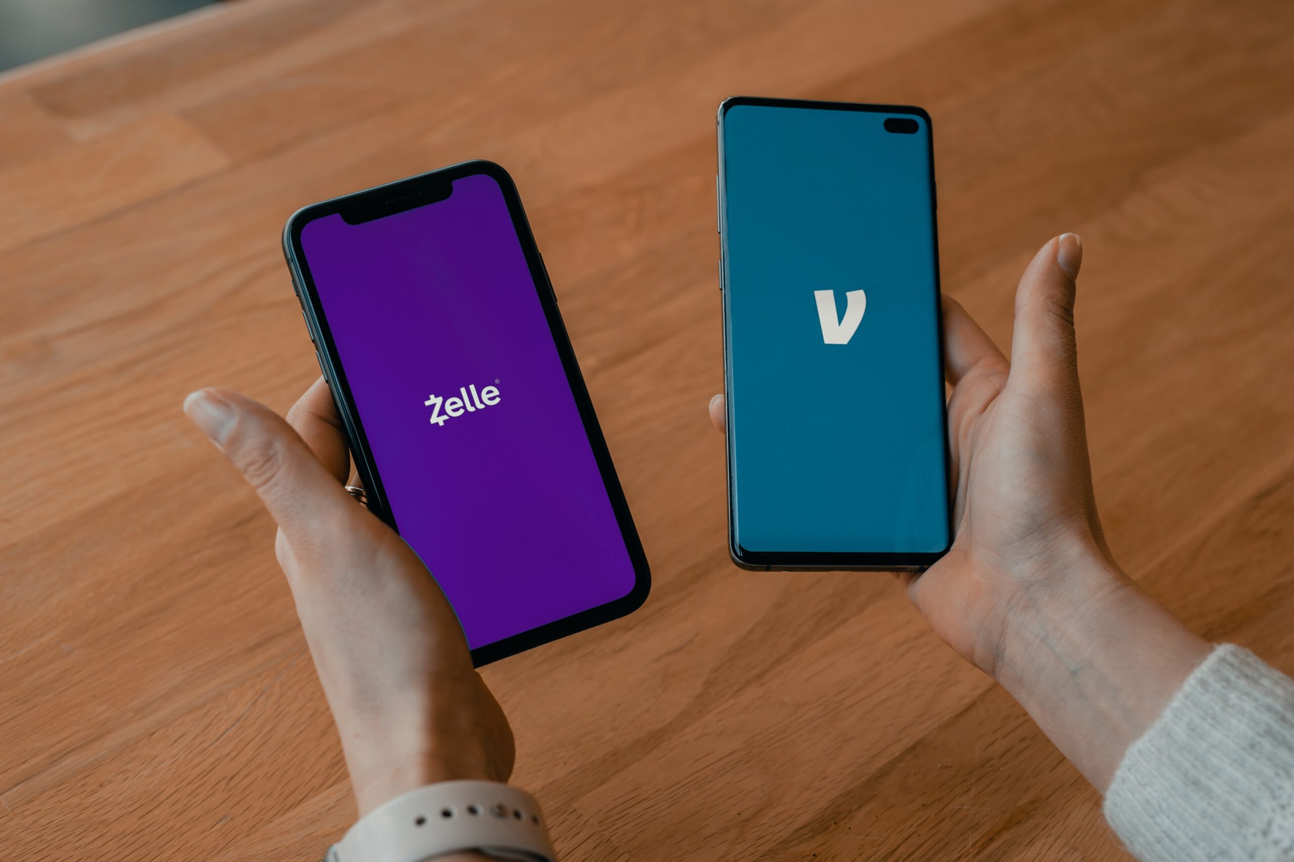 top view of a wooden desk as someone holds a phone in each hand showing p2p payments options, Zelle and Venmo